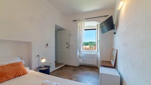 Een slaapkamer met een bed, nachtkastje met lamp, een aan de muur bevestigde televisie, raam met gordijnen en uitzicht op de omgeving.