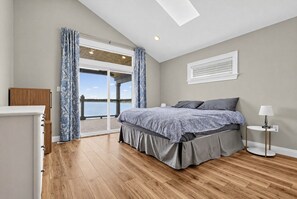 Een slaapkamer met een groot bed, een houten dressoir, een nachtkastje en uitzicht op het water door een glazen deur.