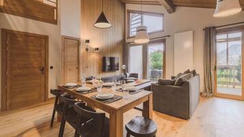 A dining area with a wooden table set for six, surrounded by chairs. There is a sofa and a television mounted on the wall.