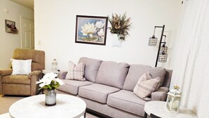 Un salón con un sofá gris, una mesita de centro redonda, una mesita auxiliar y una silla. Hay un cuadro en la pared y un jarrón con flores sobre la mesita de centro.