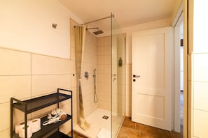 Un bany amb una cabina de dutxa de vidre, una prestatgeria de metall negre amb articles de tocador, i una porta blanca.
