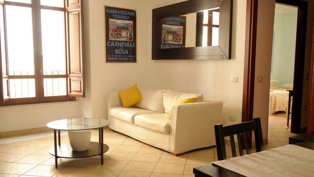 Ein Wohnzimmer mit einem weißen Sofa, einem Glastisch und einem Esstisch. An den Wänden hängen zwei Poster, und eine Tür führt in einen anderen Raum.