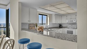 Eine moderne Küche mit weißen Schränken, einer Granit-Arbeitsplatte und einem großen Fenster. Drei blaue Barhocker und Blick auf die Stadt durch das Fenster.