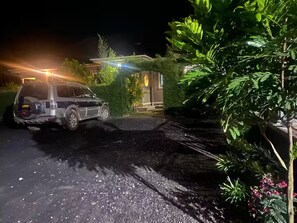 Een nachtelijke scène met een geparkeerde SUV op een oprit, omringd door groen en een huis met verlichte ramen.