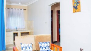 Ein Wohnzimmer mit einer Couch, zwei gemusterten Kissen, einer Küchenzeile inklusive Spüle und Schränken sowie einem Flur, der zu einem weiteren Raum führt.