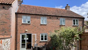 A two-story brick house with a tiled roof, a patio with outdoor furniture, and a garden with various plants. - Wild Strawberry Cottage is a charming hideaway within a peaceful courtyard (Burnham Market, Norfolk)
