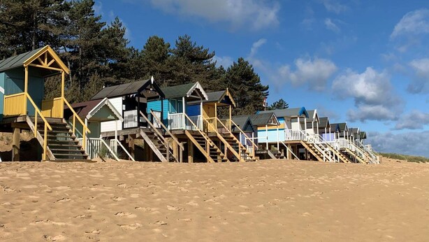 A row of beach huts with wooden stairs leading up to them, set against a sandy beach and trees in the background. - Traditional flint cottage, tucked away in the town of Wells-next-the-Sea (Wells-next-the-Sea)