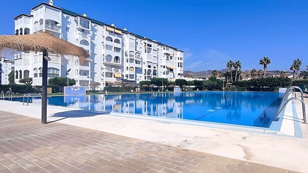 Ein Schwimmbecken mit klar blauem Wasser, umgeben von einer gefliesten Liegefläche. Im Hintergrund ein mehrstöckiges Gebäude mit Balkonen und einem reetgedeckten Aufbau.