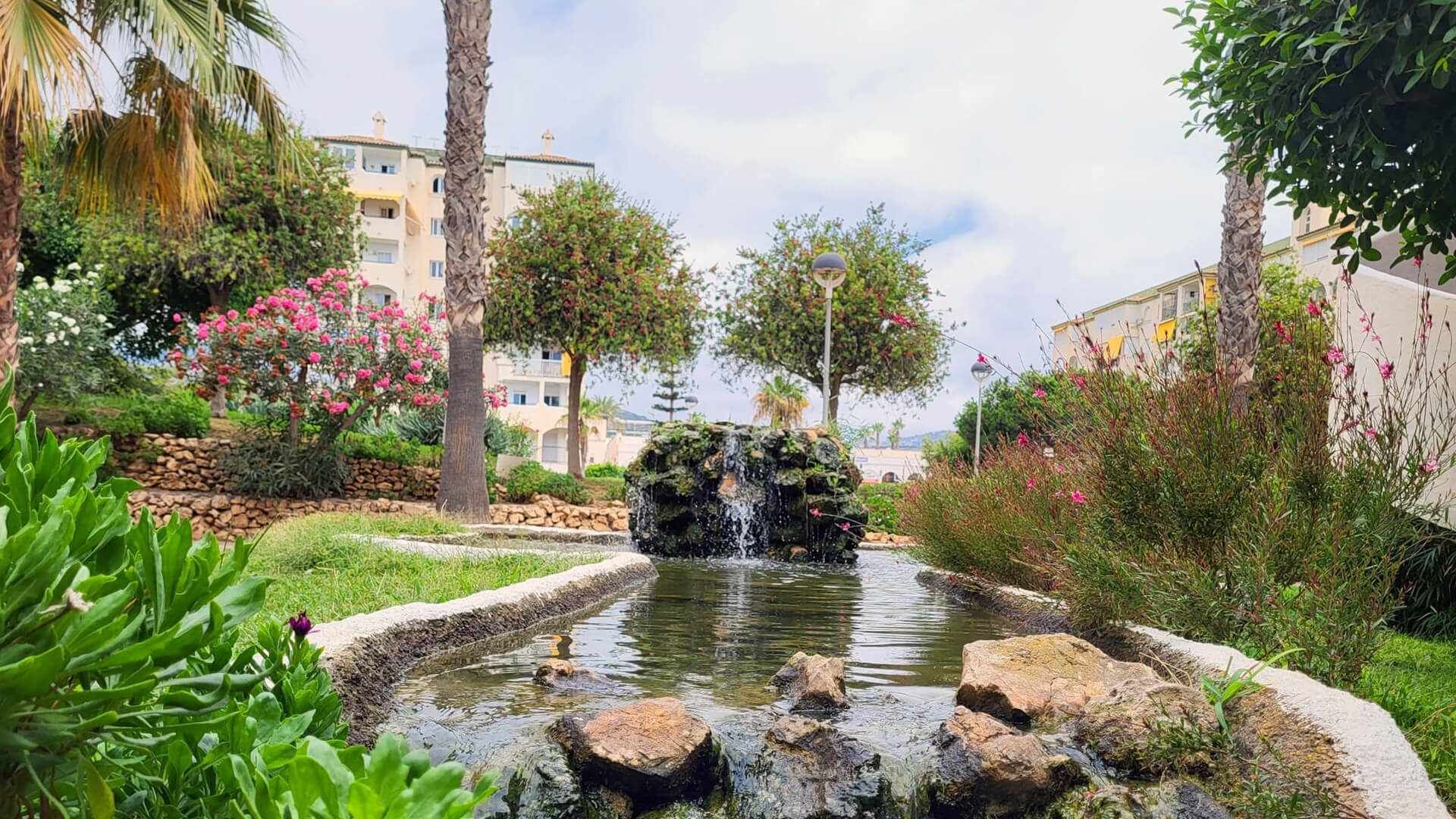Jardí paisatjístic amb una petita cascada, envoltat de vegetació exuberant i plantes florentes.