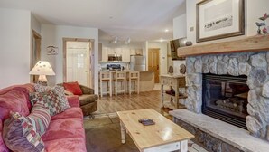 A living room with a sofa, coffee table, fireplace, kitchen in the background, and a framed picture on the wall. - Spacious 3BR Ski Condo | Balcony, Pool & Fireplace (Keystone)