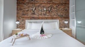 A hotel room with a brick wall, a large bed with white bedding, two bedside tables with lamps, and a small wooden side table with a plant and a glass. - Charming Family Escape in Trendy Plateau Montreal (Montréal)