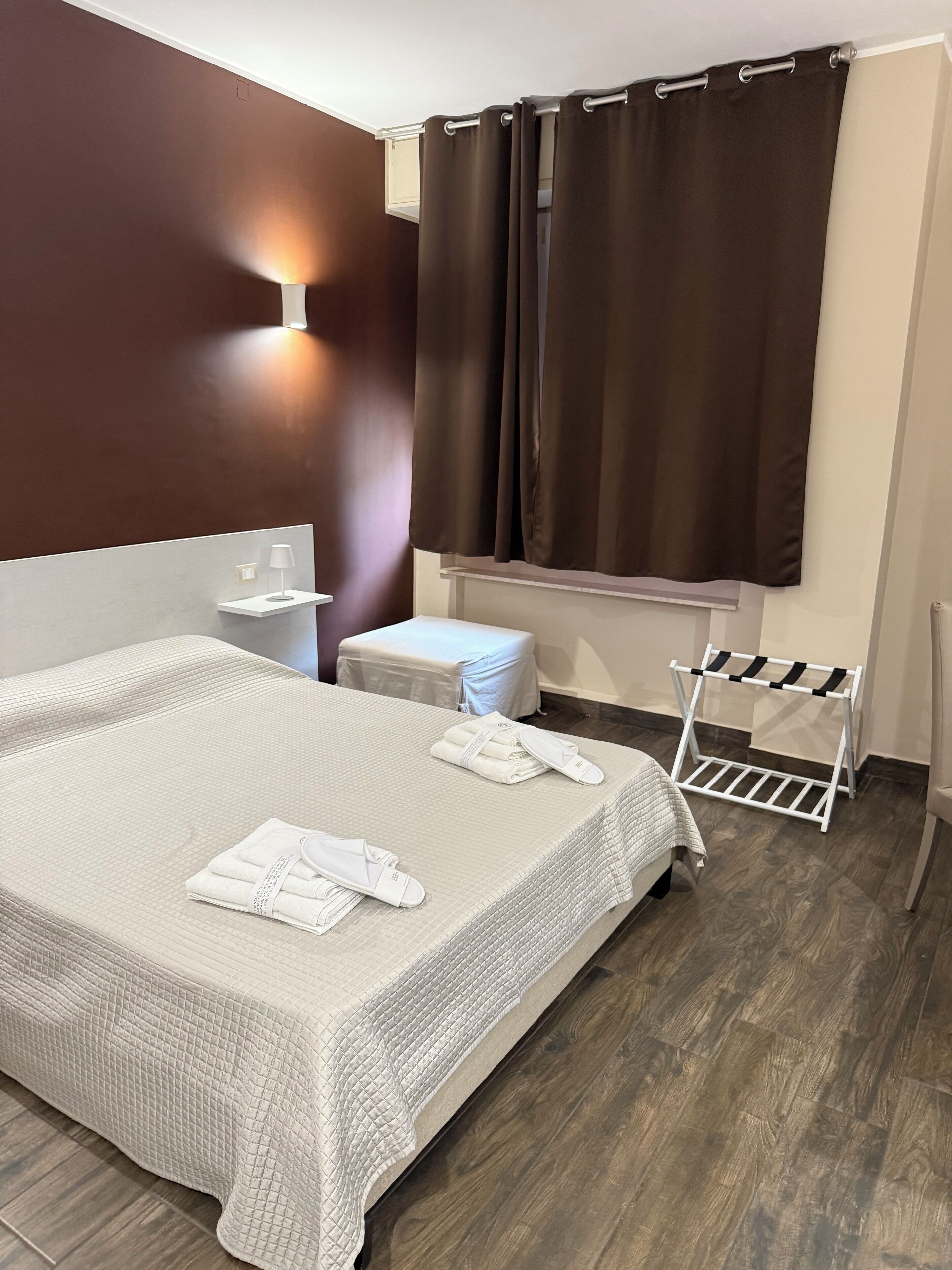 Camera d'albergo con un letto grande, un comodino, uno sgabello, un portasacchi e una finestra con tende.