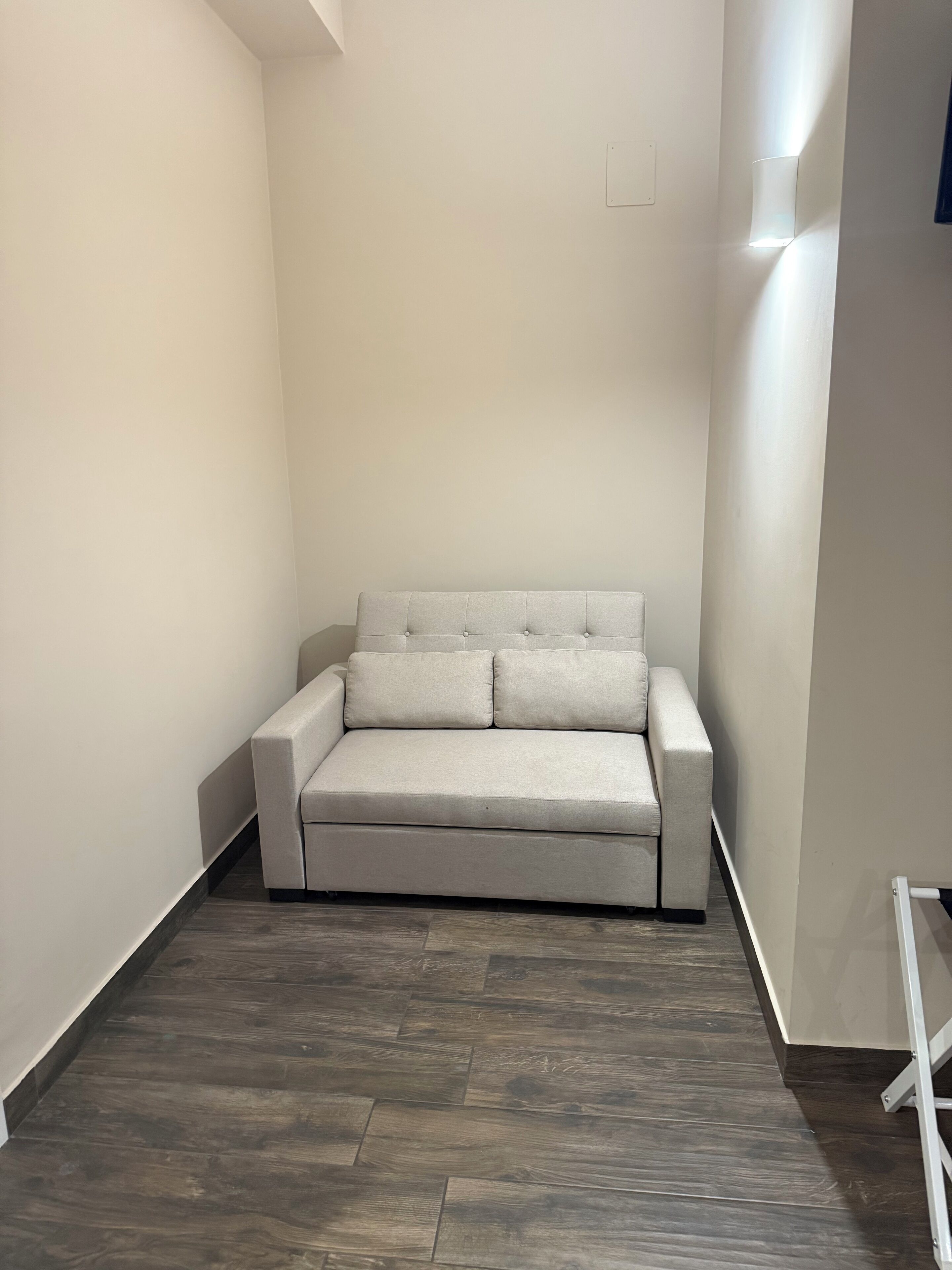 A small, neutral-colored sofa in a corner with a wooden floor and white walls.