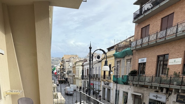 A balcony with wicker furniture, a tiled floor, and a view of a city street with buildings and shops. - B&B Leus Appartment (Reggio Calabria)