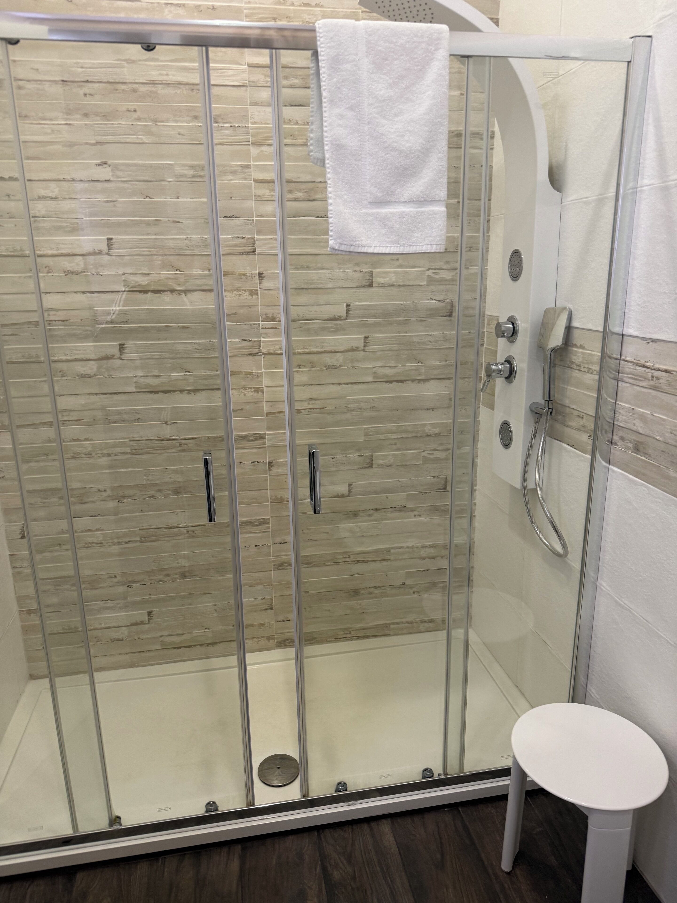 Een douchecabine met glazen wanden, een houten paneelwand en een witte handdoek die aan de deur hangt, plus een klein wit krukje.