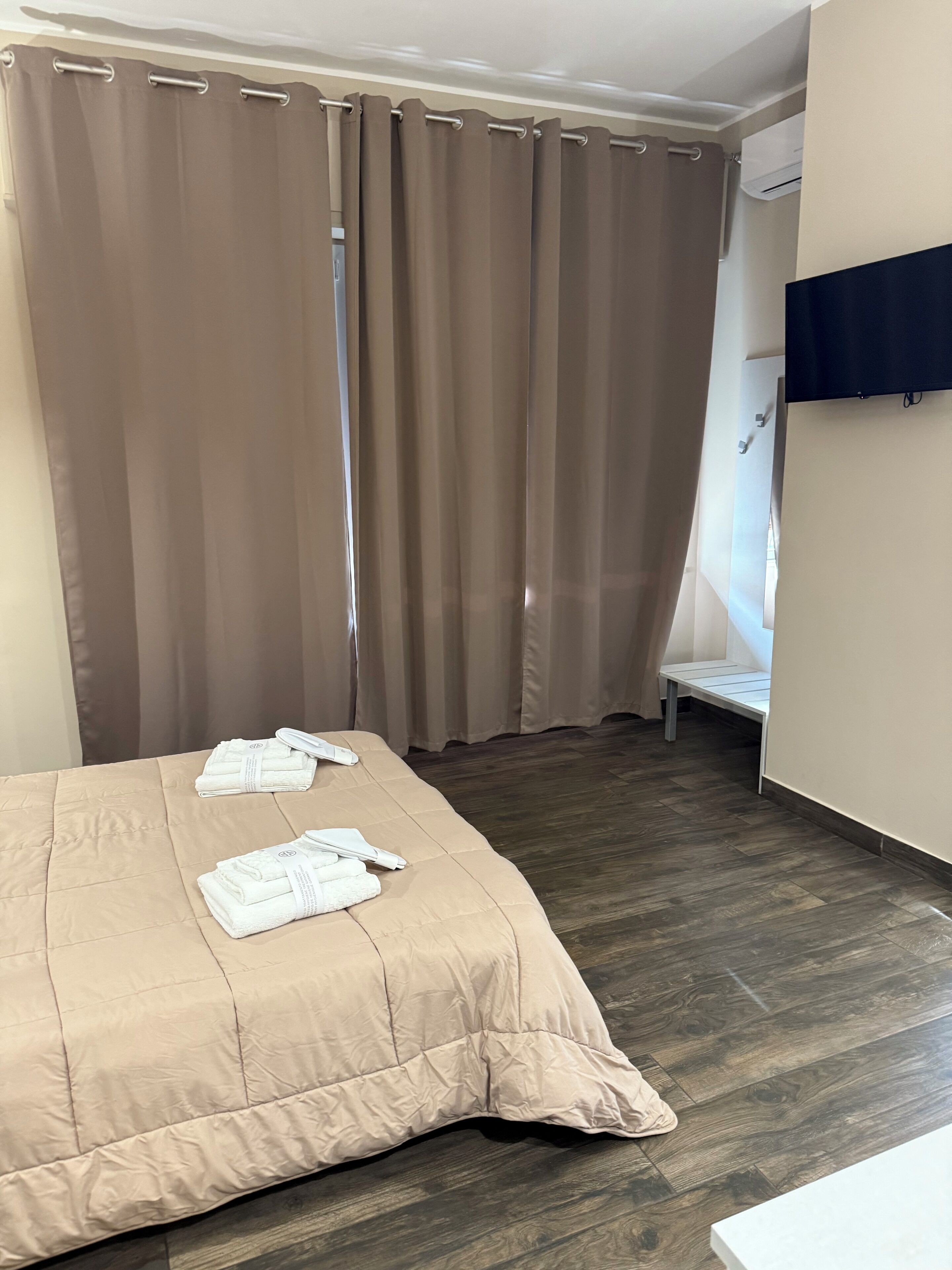 A bedroom with a bed, beige curtains, a wooden floor, and a white wall.