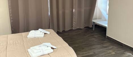 A bedroom with a bed, beige curtains, a wooden floor, and a white wall.