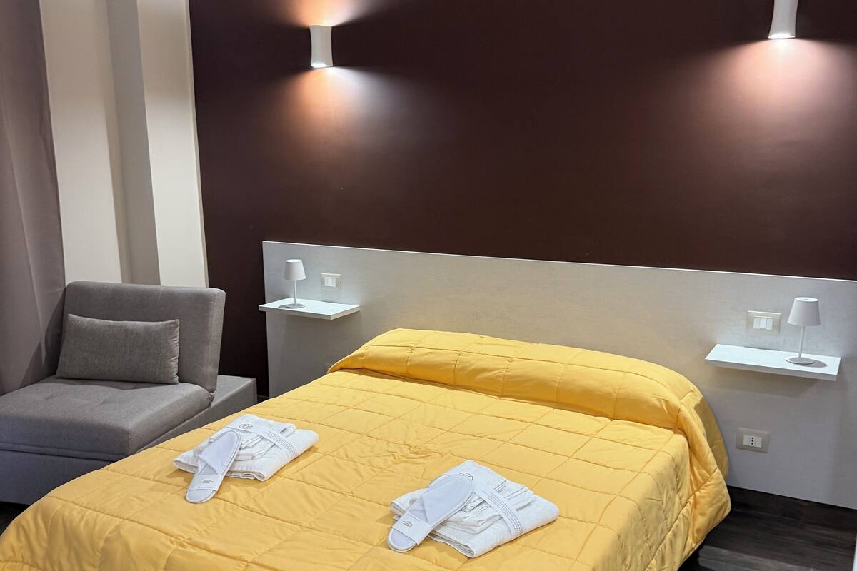 Habitación de hotel con una cama amarilla, sofá gris y dos lámparas fijadas a la pared.