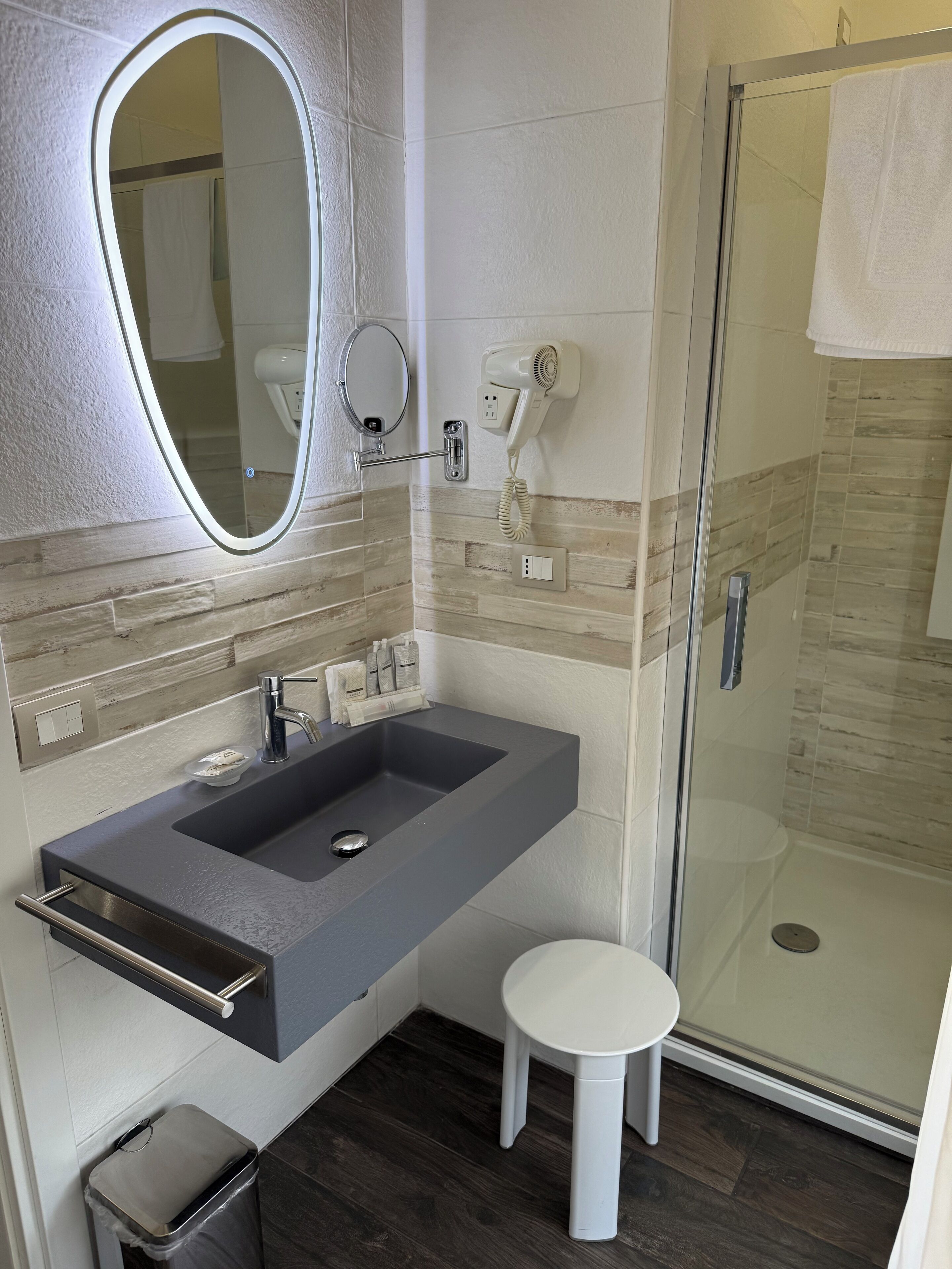 A modern bathroom with a large mirror, a built-in sink, a handheld showerhead, and a small stool.