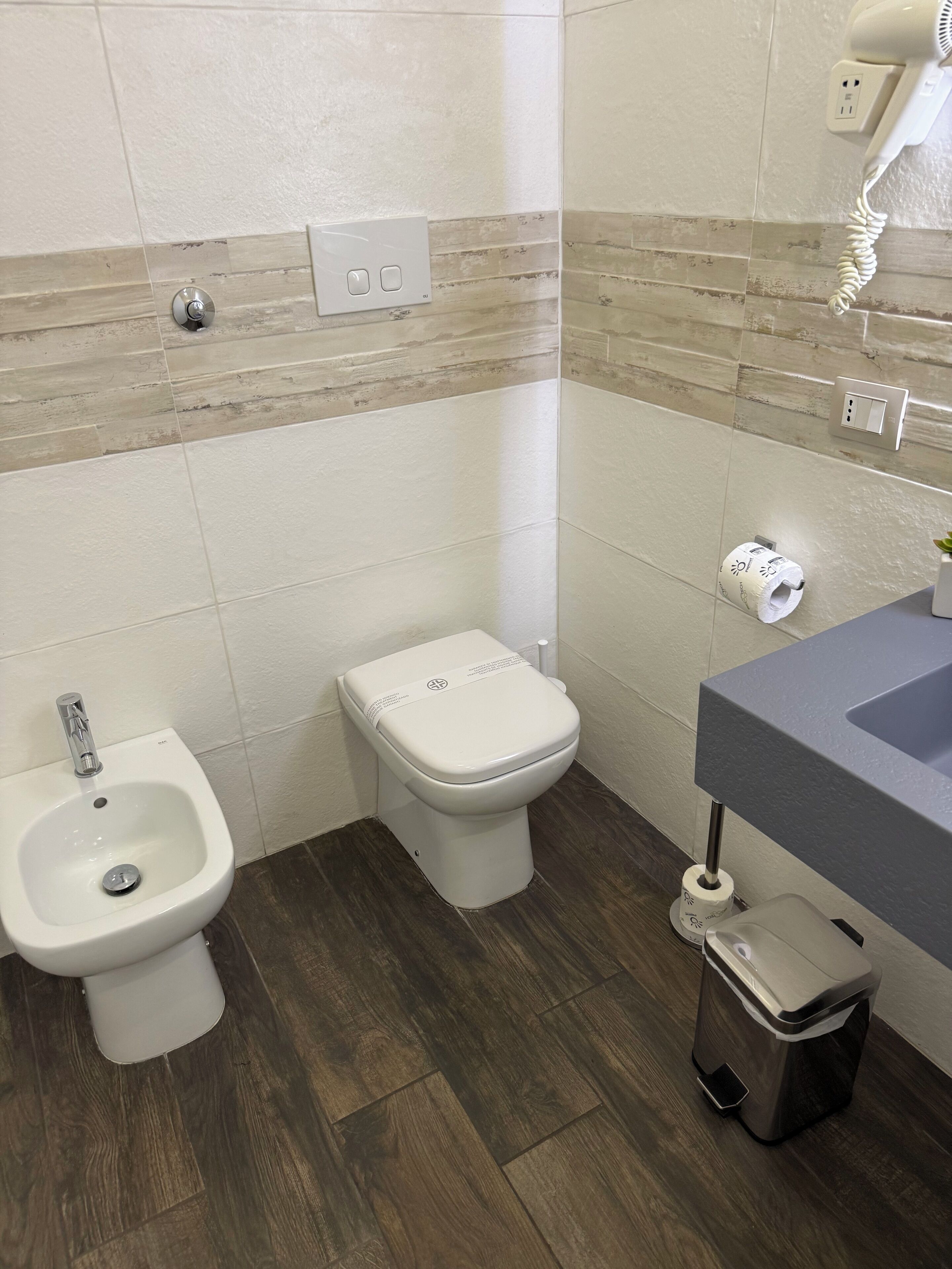 Un baño con bidé, inodoro, lavamanos y un secador de pelo en la pared.