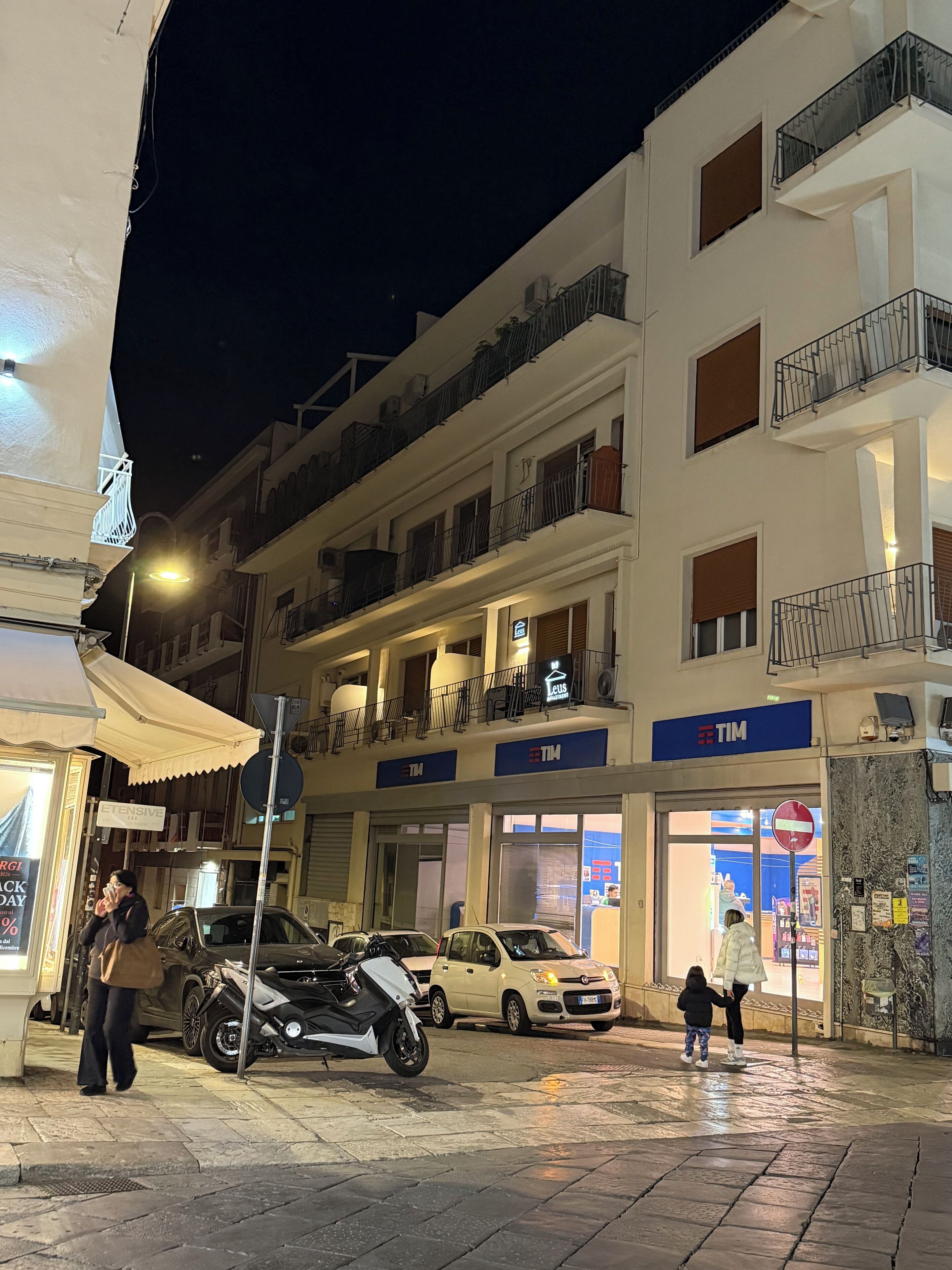Un edificio de varios pisos con balcones, una tienda TIM y automóviles estacionados.
