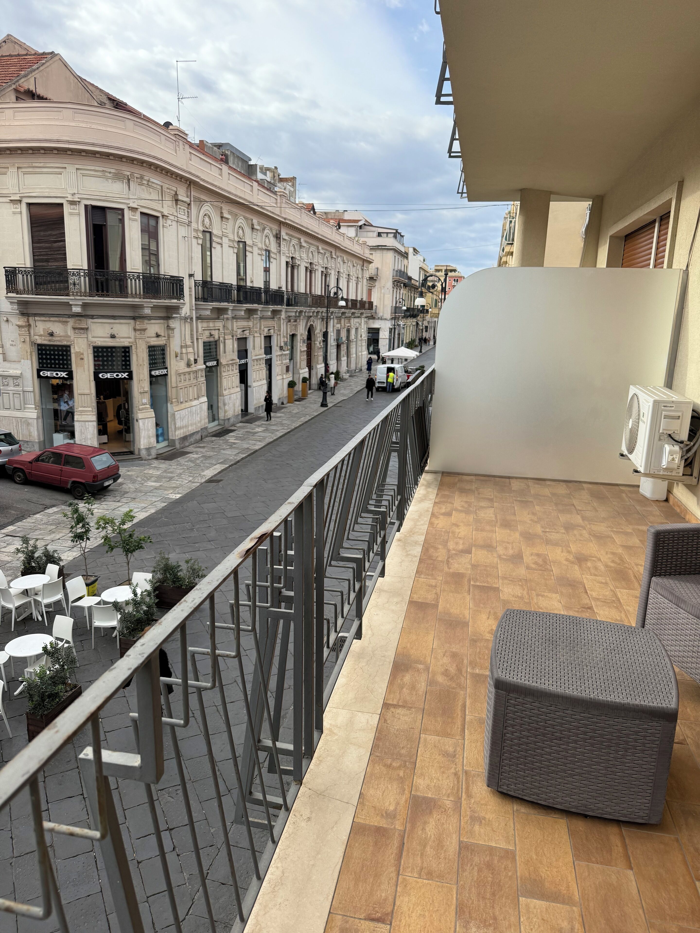 A balcony with wicker furniture, a tiled floor, and a street view with buildings and a red car.