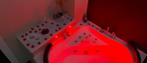 A modern bathroom with a large, illuminated bathtub, a small table with rose petals and a heart-shaped dessert, and a decorative wall piece.