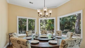 Uma sala de jantar com uma mesa redonda, cadeiras com estampas vintage, um candelabro e grandes janelas com vista para o verde.