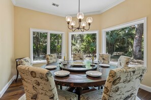 A dining area with a round table, chairs with vintage patterns, a chandelier, and large windows with a view of greenery.