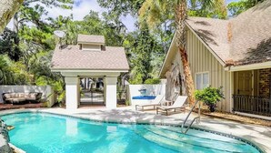 Uma piscina com um pequeno gazebo, cadeiras de descanso e uma casa com telhado em azulejos.