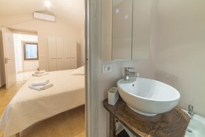A hotel room with a bed, a large mirror, a white sink, and a wooden vanity. - S'Iscola (Bosa)