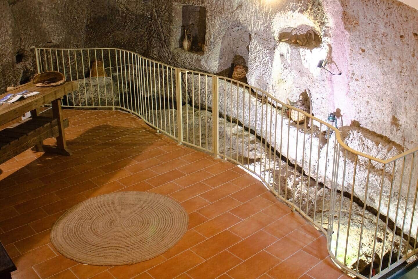 Un interior amb parets de pedra, un terra de rajola, una banca de fusta i una barana de metall.