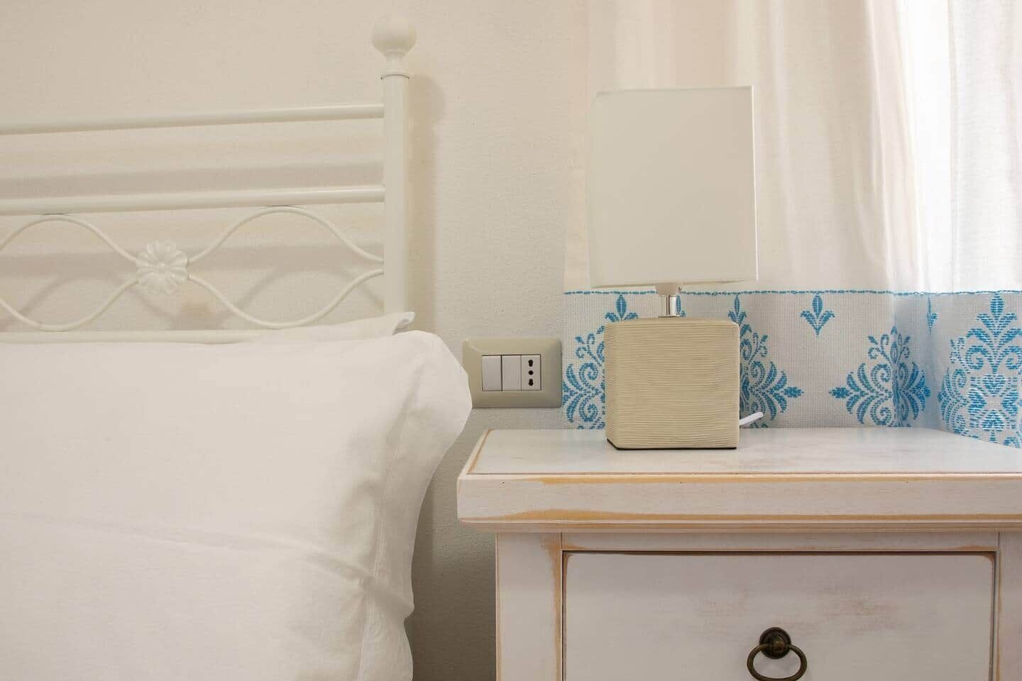 Un lit blanc avec une tête de lit en métal, une table de chevet avec une lampe et un rideau à motifs.