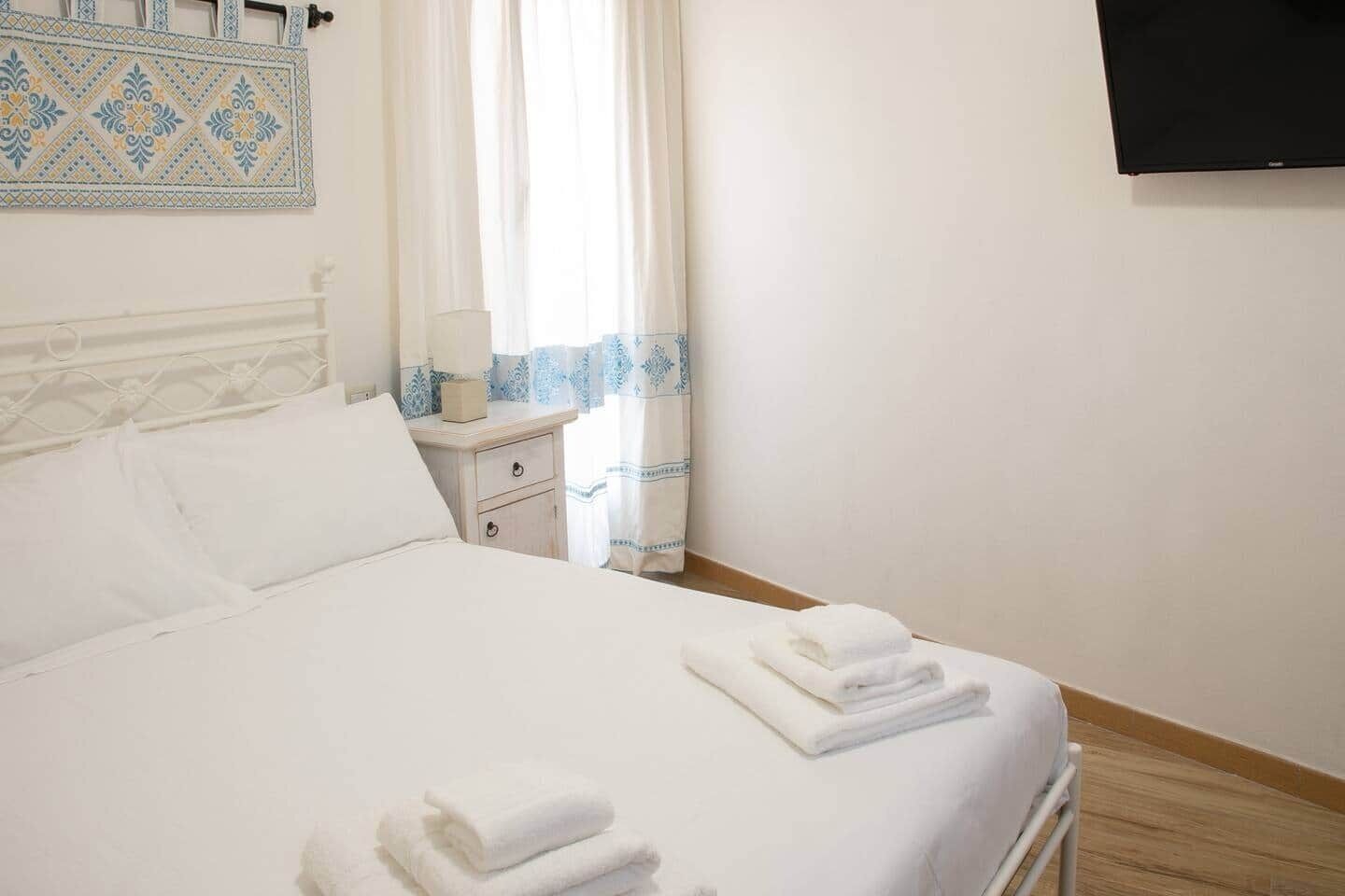 A neatly made bed with white linens and folded towels, a bedside table with a lamp, and a wall-mounted TV.