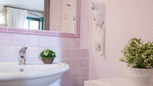 Un bagno con un lavandino bianco, una piccola pianta in vaso, un asciugacapelli montato sulla parete e uno specchio sopra il lavandino.