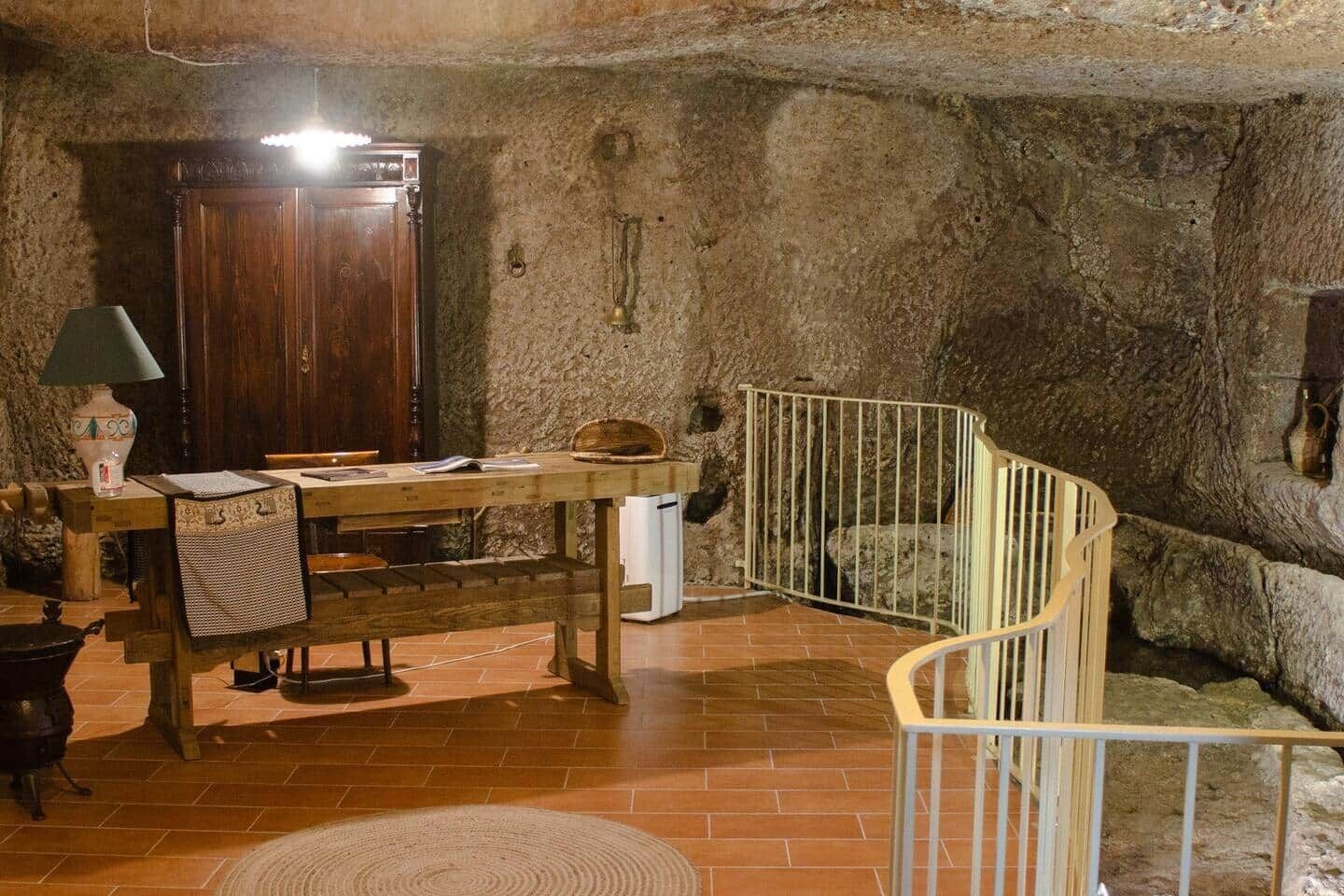 A rustic room with a wooden table, a lamp, and a chair. There is a staircase with a railing leading to a higher level.
