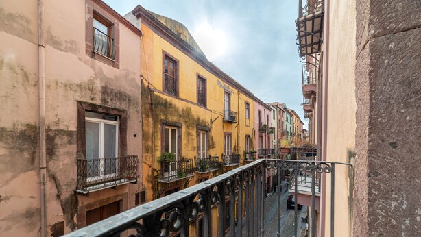 Un carrer estret amb edificis a ambdós costats, balcons amb baranes i un cel clar.