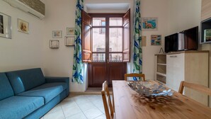 Un saló amb un sofà, una taula de dinar de fusta, cadires, un televisor i una finestra amb cortines.