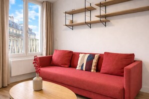 Un sofá rojo con un jarrón de bayas rojas, una mesa de madera y una ventana con vistas a edificios.