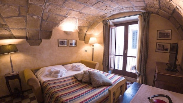 A cozy room with a checkered floor, a bed with striped bedding, a red kettle, and a window with curtains. - Sardinian Gallery Vittorio (Bosa)