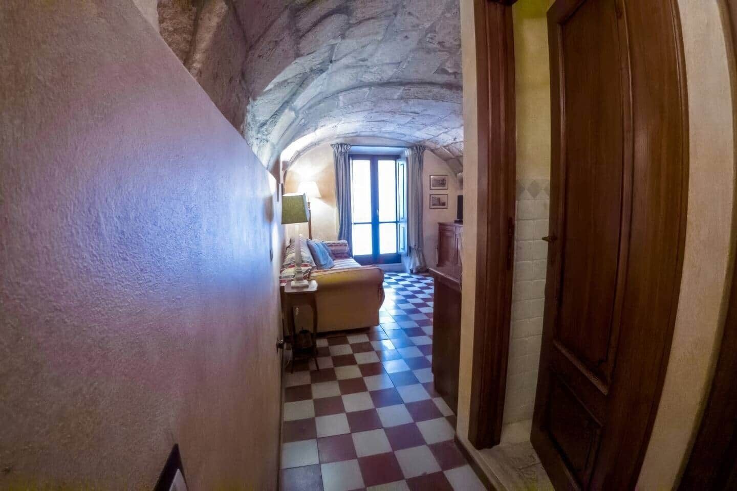 Un couloir étroit, dont le sol est recouvert de carreaux, mène à une pièce où se trouvent un lit et une fenêtre.