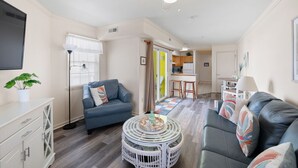 A compact living space with a sofa, a chair, a small table, a television, and a kitchenette. - Surfrider F — 70 Steps to the Sand • 3BR/2BA • 2 Balconies • Sleeps 8 (Dewey Beach)
