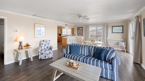 A living room with a striped sofa, a coffee table, a chair, and a dining table with chairs. There is a ceiling fan, a kitchen in the background, and a window with curtains. - 70 Steps to the Sand • 2BR/1BA • 2 Balconies • Sleeps 6 (Dewey Beach)