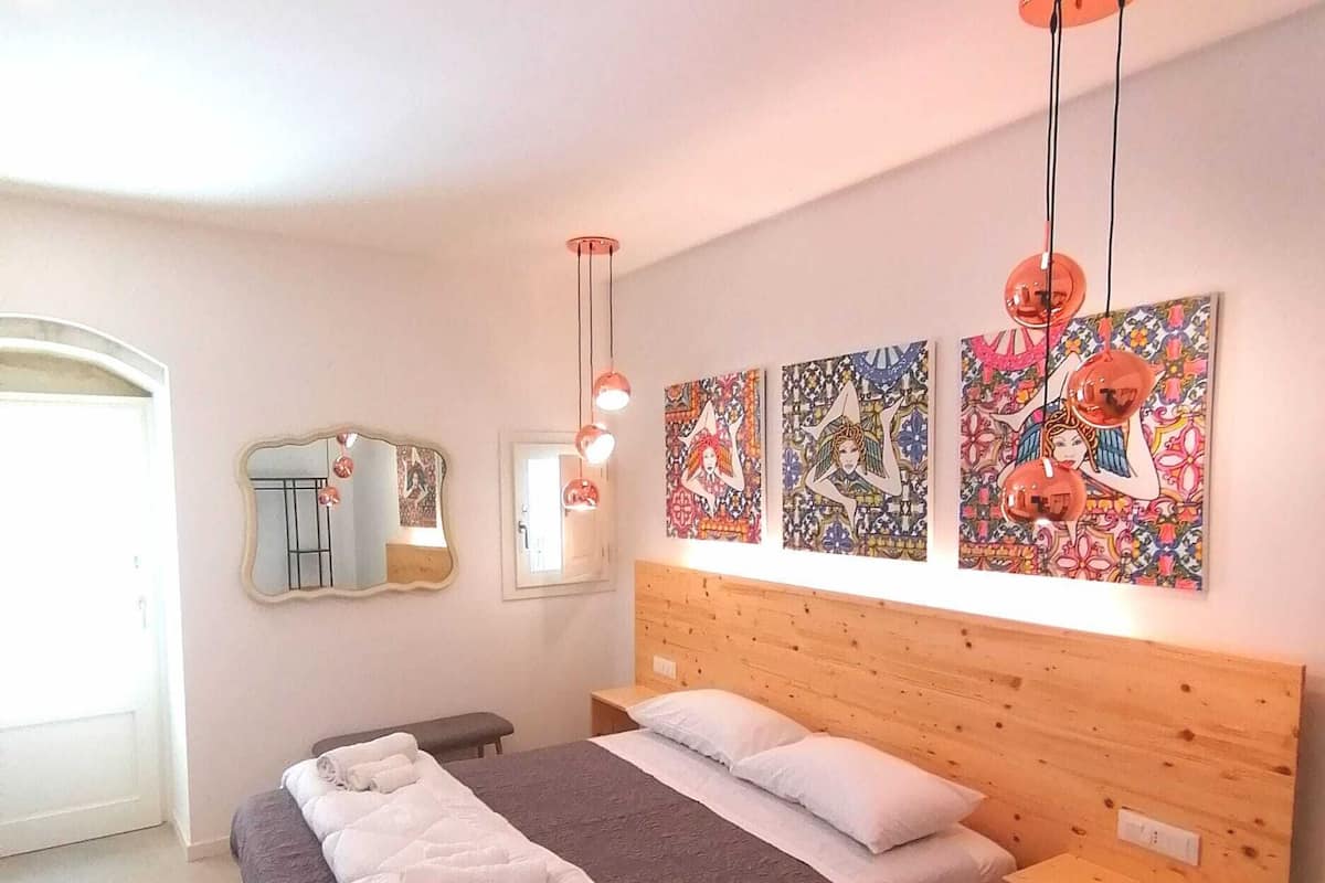 Ett sovrum med en sänggavel i trä, en säng med vita och grå sängkläder, en bänk och en spegel. Det finns tre konstverk som hänger på väggen och två hänglampor.
