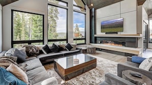 A modern living room with a large TV, fireplace, and large windows offering a forest view. - Walk-to-Ski Retreat | Serac Lodge (Breckenridge)