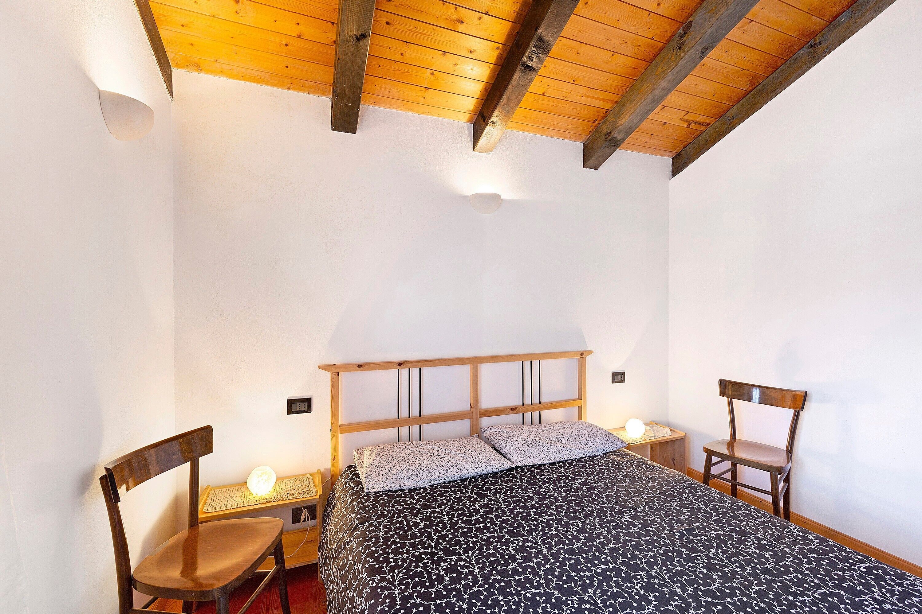 Un dormitorio con techo de madera, una cama con colcha estampada, dos sillas y lámparas de noche.