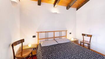 Un dormitorio con techo de madera, una cama con colcha estampada, dos sillas y lámparas de noche.