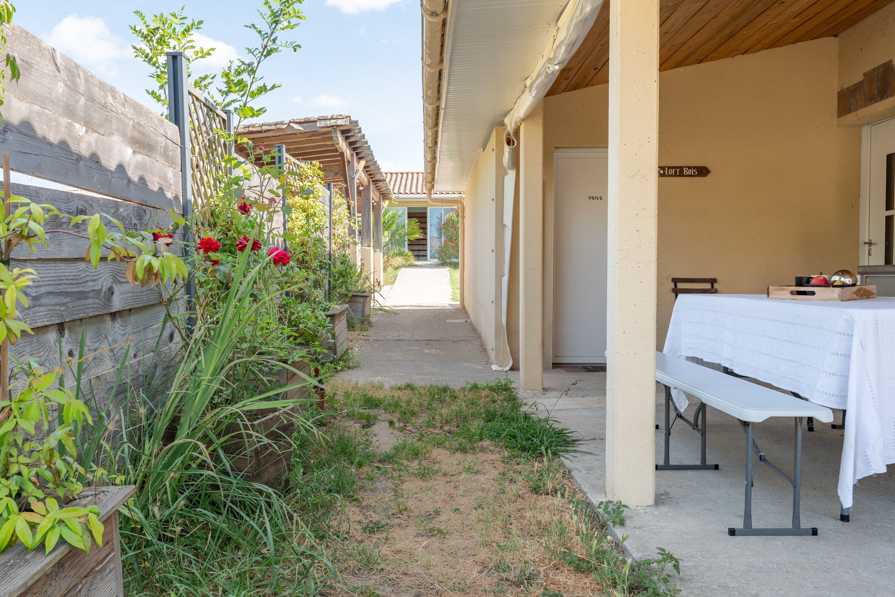 A covered outdoor area with a table set up, a wooden fence, and a pathway leading to a building.
