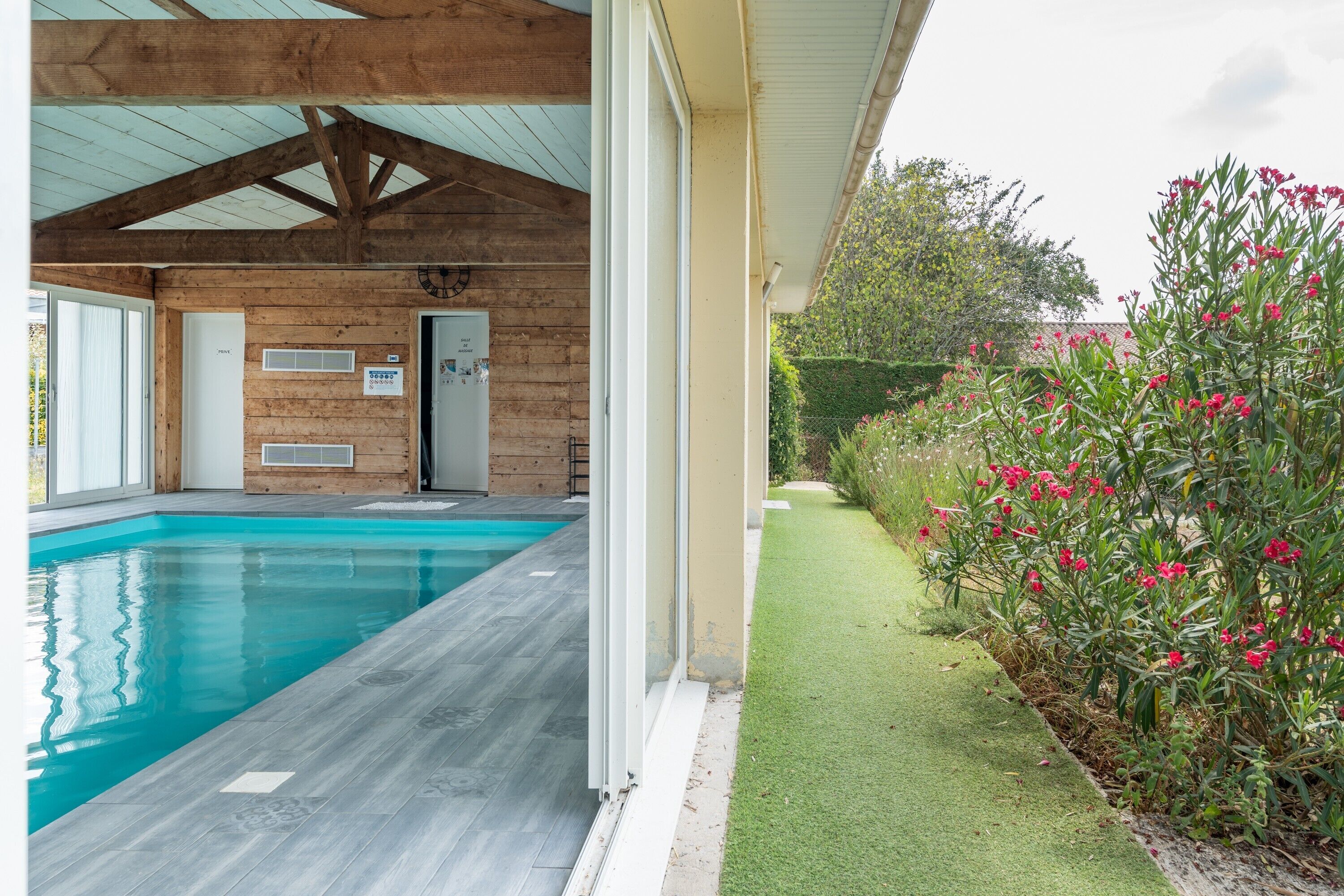 A swimming pool with a wooden deck, a covered area with a sloped roof, and a well-maintained lawn with flowering plants.