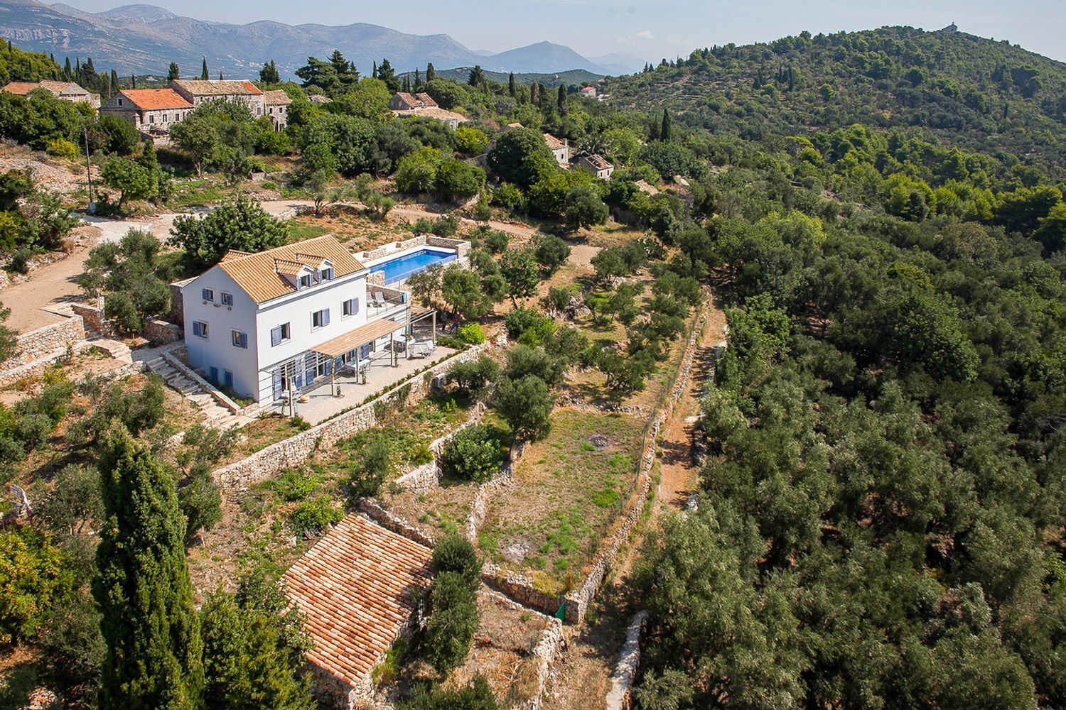 A large white building with a swimming pool, surrounded by trees and hills.
